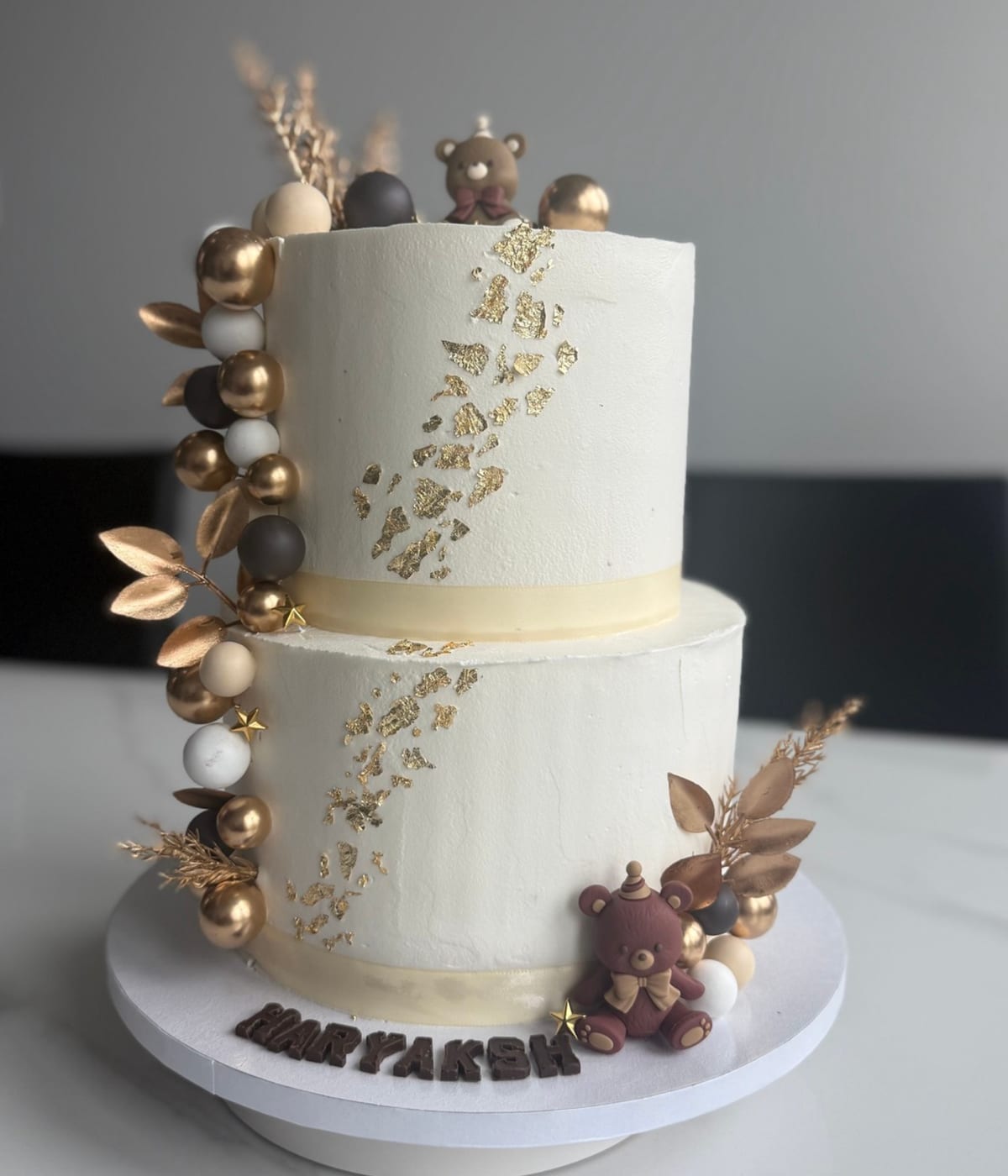 Two-tier ivory buttercream cake with flecks of gold leaf running diagonally, a cluster of gold, white and grey spheres up one side, dried palm leaves and two small fondant teddy bears.