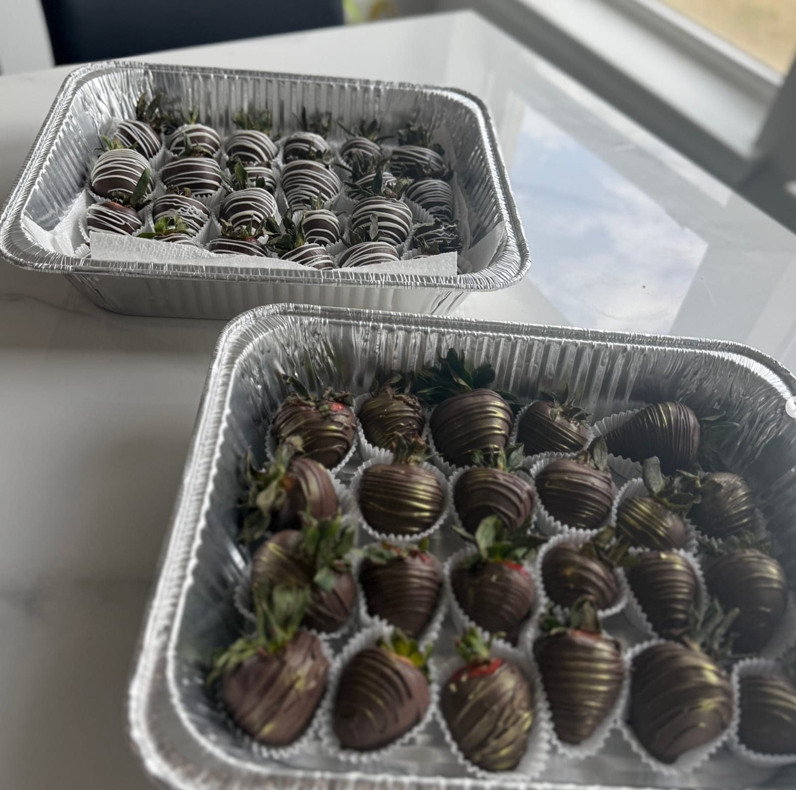 Two foil trays of whole strawberries dipped in dark and milk chocolate, each finished with a contrasting drizzle, on a kitchen counter beside a window.