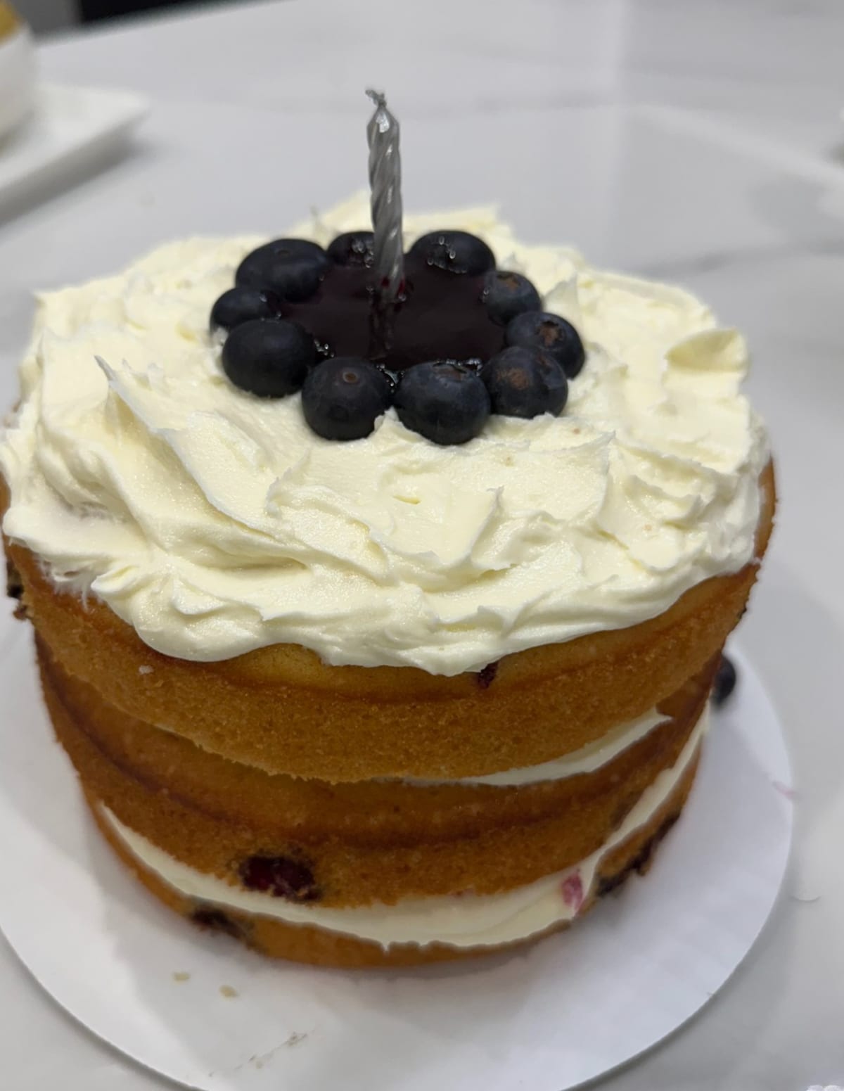 Rustic bare-sided layer cake with the sponge and cream layers showing, thick whipped cream on top and a ring of fresh blueberries around a single candle.