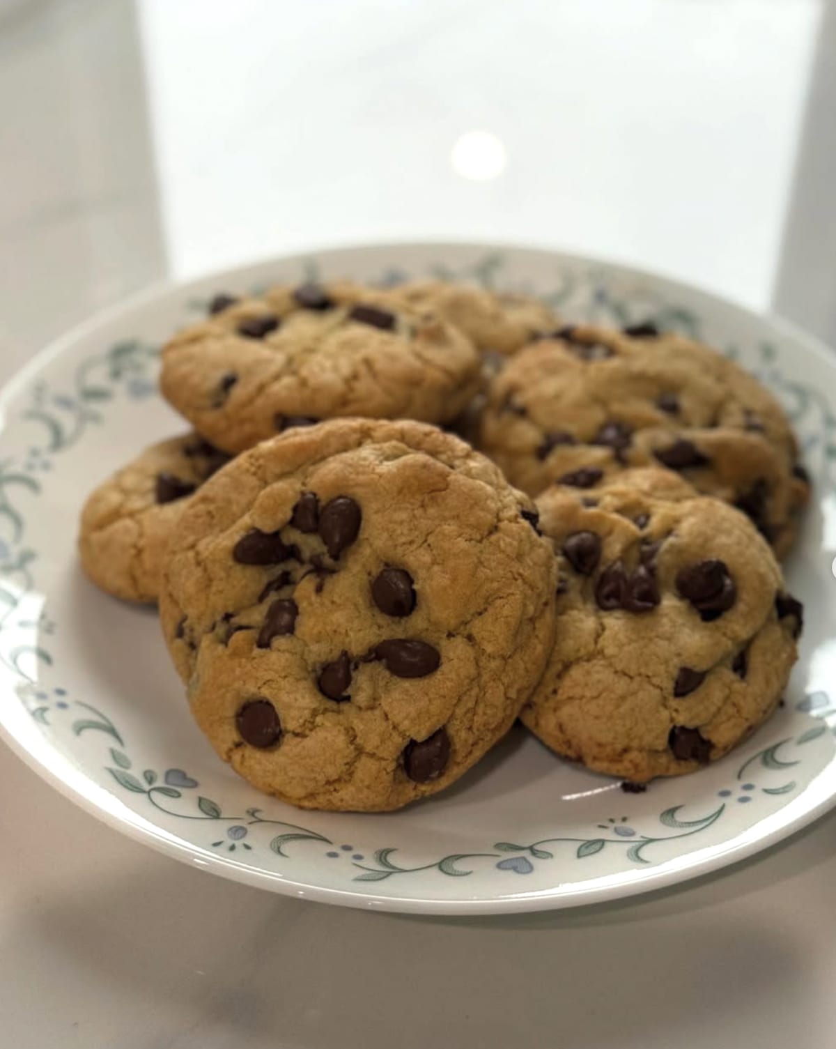 Six thick chocolate chip cookies piled on a white plate with a green floral rim.
