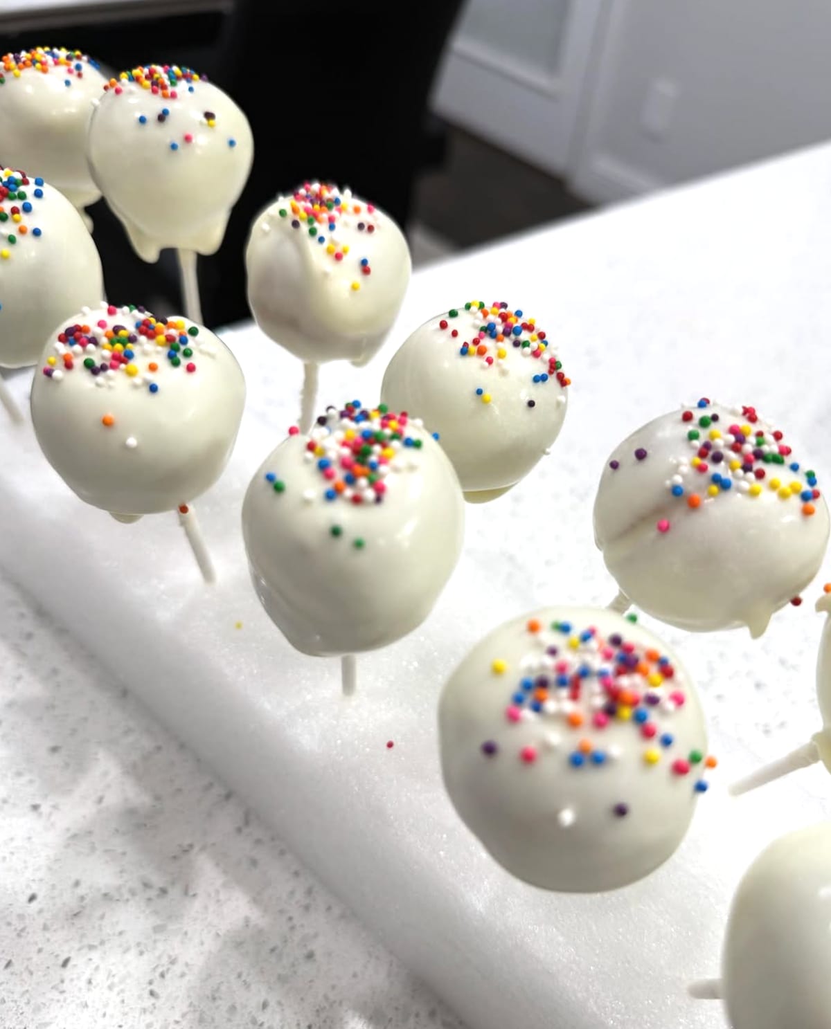 A dozen cake pops coated in white chocolate and scattered with rainbow nonpareil sprinkles, standing on sticks in a white block.
