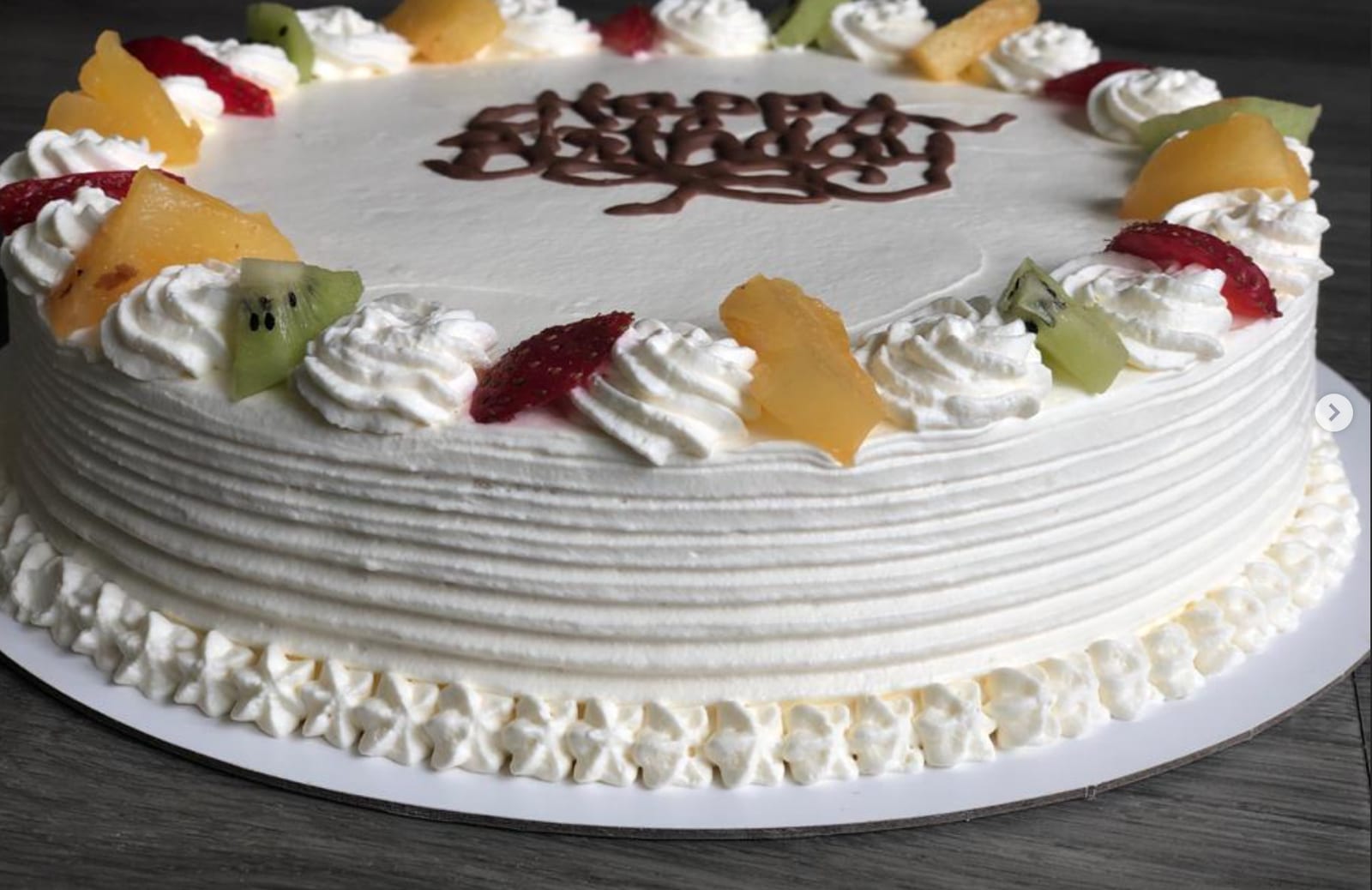 Round whipped cream cake with piped rosettes and a ring of kiwi, strawberry and peach around the edge, and a birthday message written across the top in chocolate.