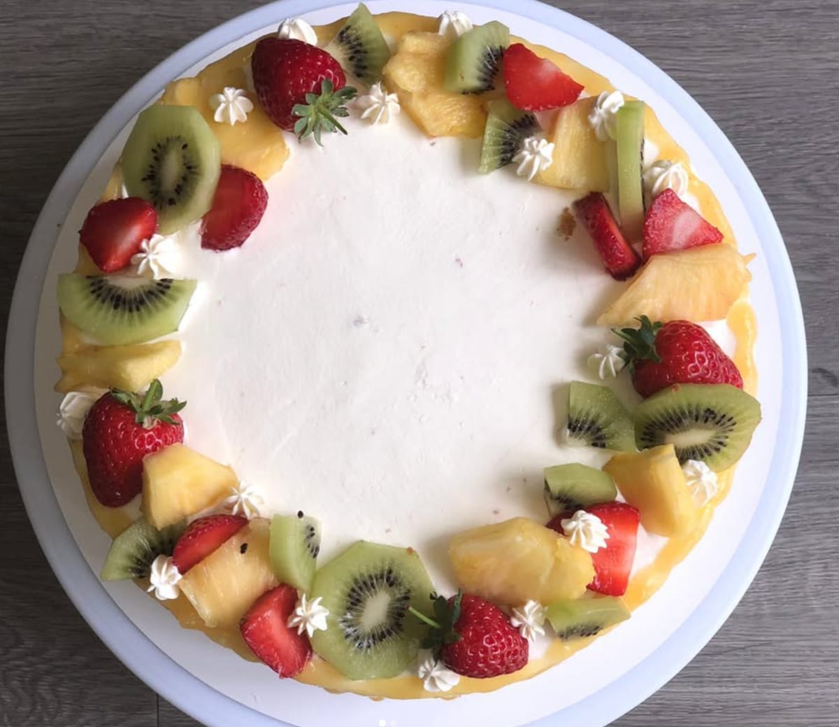 Overhead view of a round cream-frosted cake edged with a ring of sliced kiwi, strawberries and pineapple alternating with piped cream rosettes.