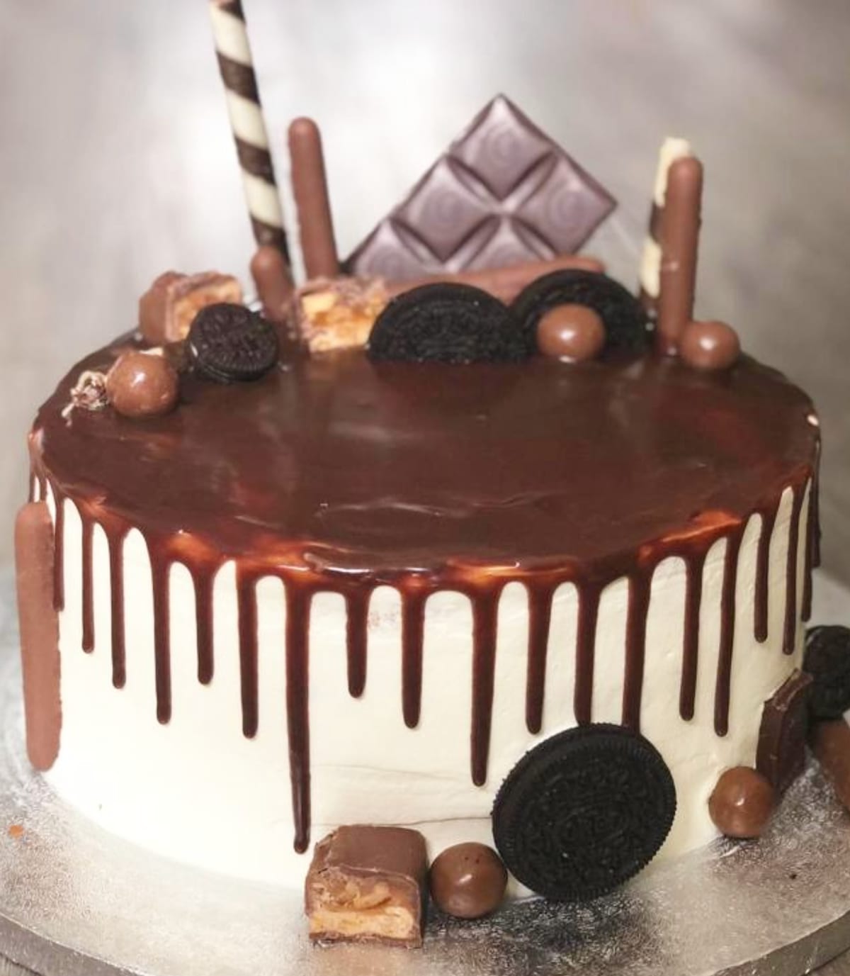 Cake with dark chocolate ganache pooled on top and dripping down white buttercream sides, loaded with Oreos, chocolate bars, malt balls and a striped wafer straw.