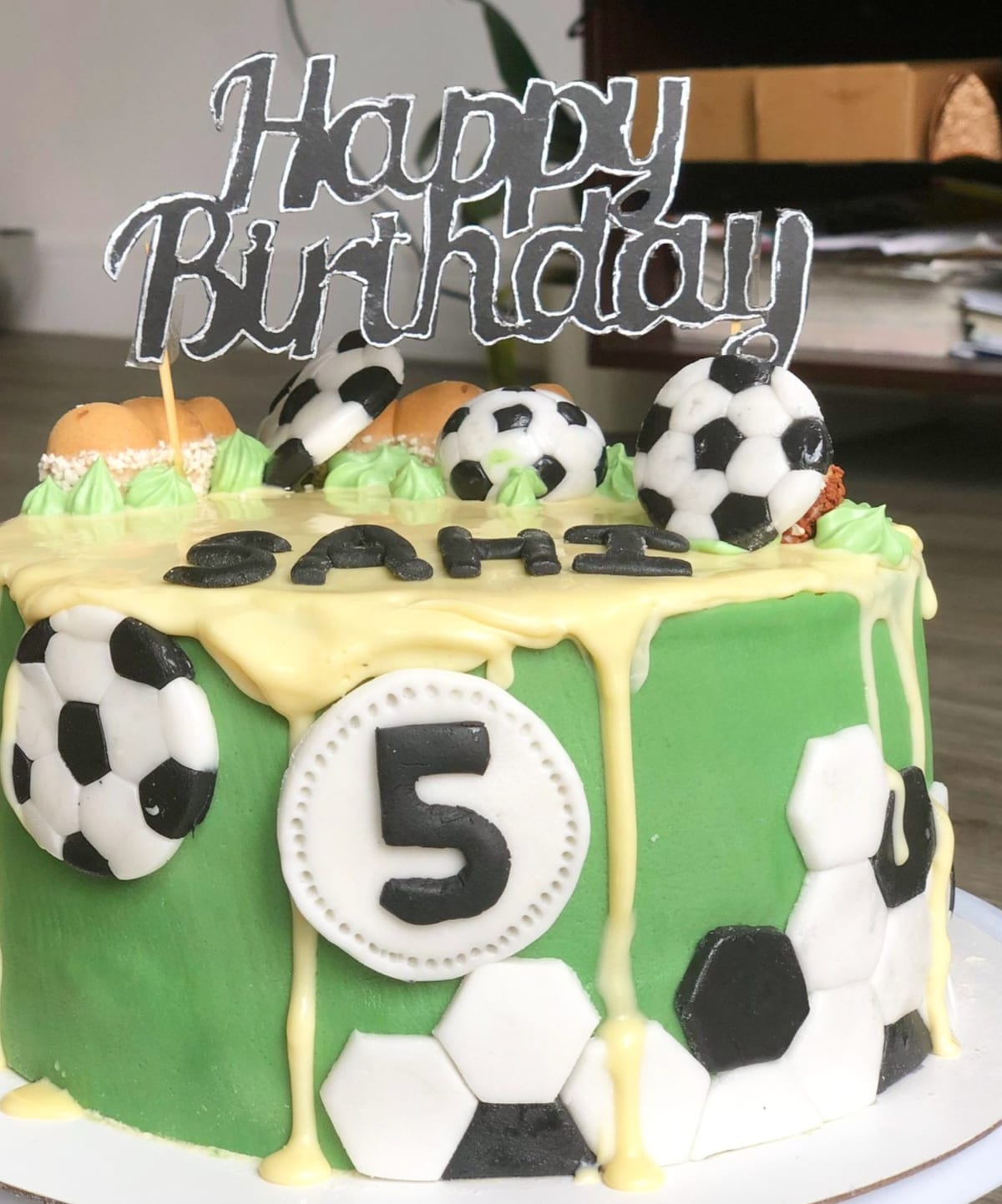 Round cake covered in green fondant with a pale drip down the sides, decorated with black and white fondant footballs, a number five medallion and a mirrored Happy Birthday topper.