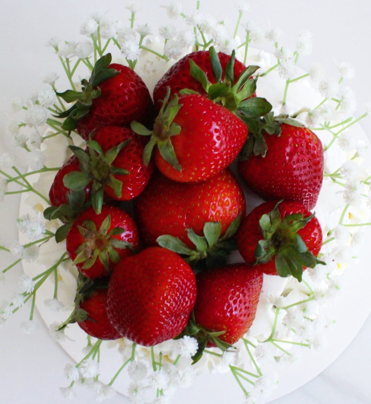 Overhead view of whole strawberries heaped on top of a white cake, surrounded by a ring of small white blossom.