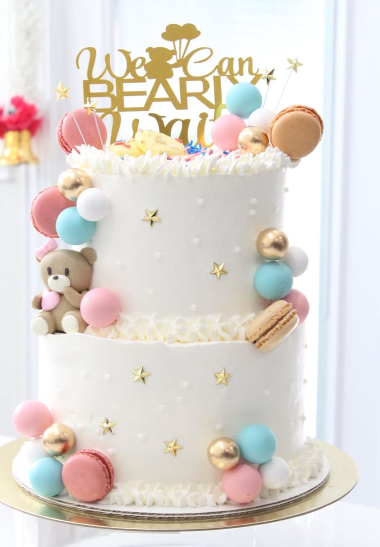 Two-tier white cake dotted with small gold stars, trimmed with pink, blue and gold spheres and macarons, with a fondant teddy bear seated on the lower tier and a gold baby shower topper on top.